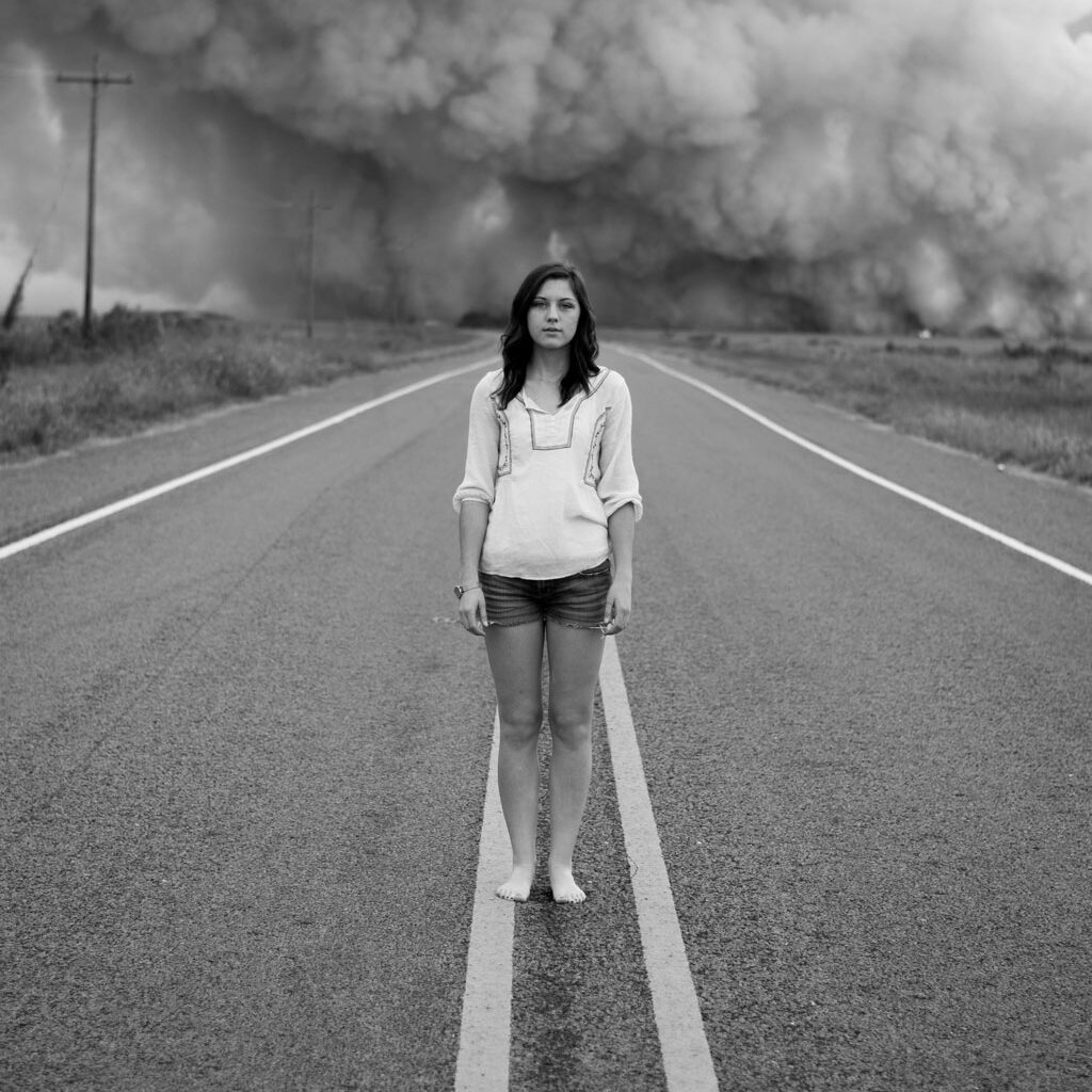 black and white photo of woman standing on road's divider with dust clouds in background